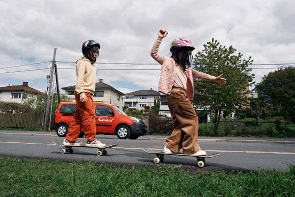 To barn på skateboard på siden av en vei. En rød bil fra Posten i bakgrunnen.