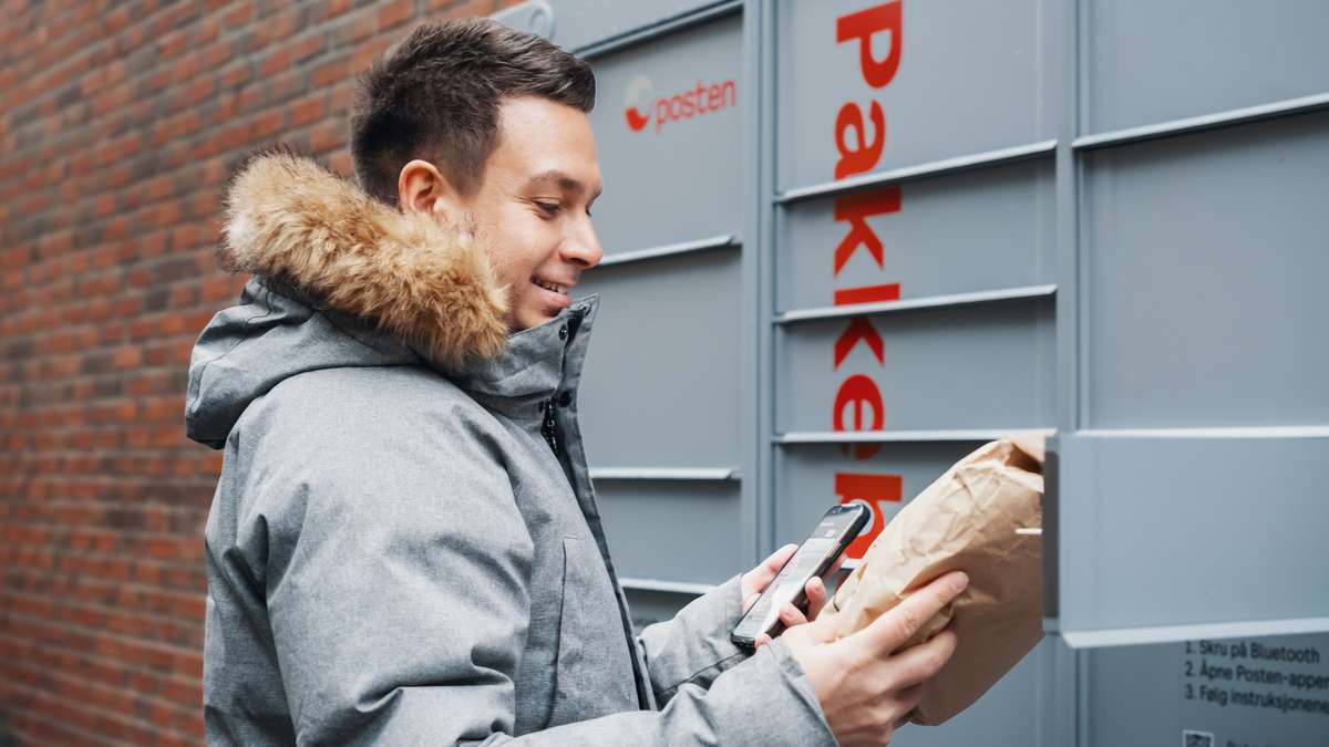 Mann med vinterjakke står med pakke og mobil ved en av Postens pakkebokser