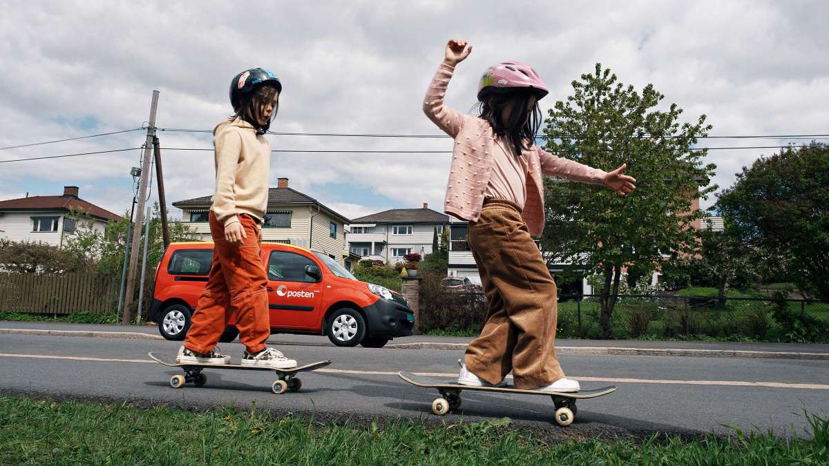 To barn på skateboard på siden av en vei. En rød bil fra Posten i bakgrunnen.