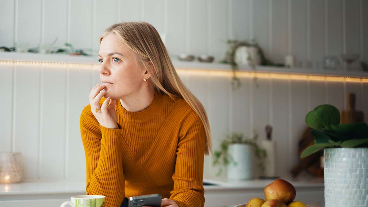 Kvinne i tenkende positur på et kjøkken, med mobil i hånden