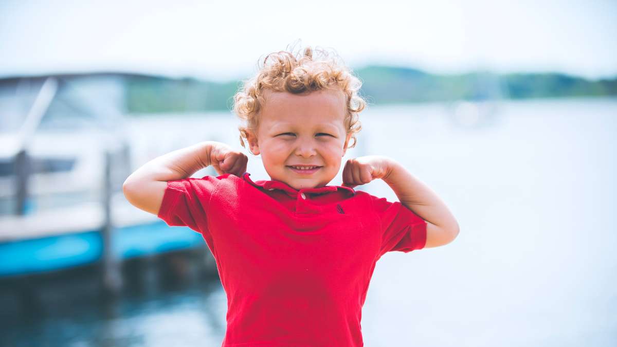 Liten gutt med krøller og rød trøye, som står foran et vann og viser at han er sterk.