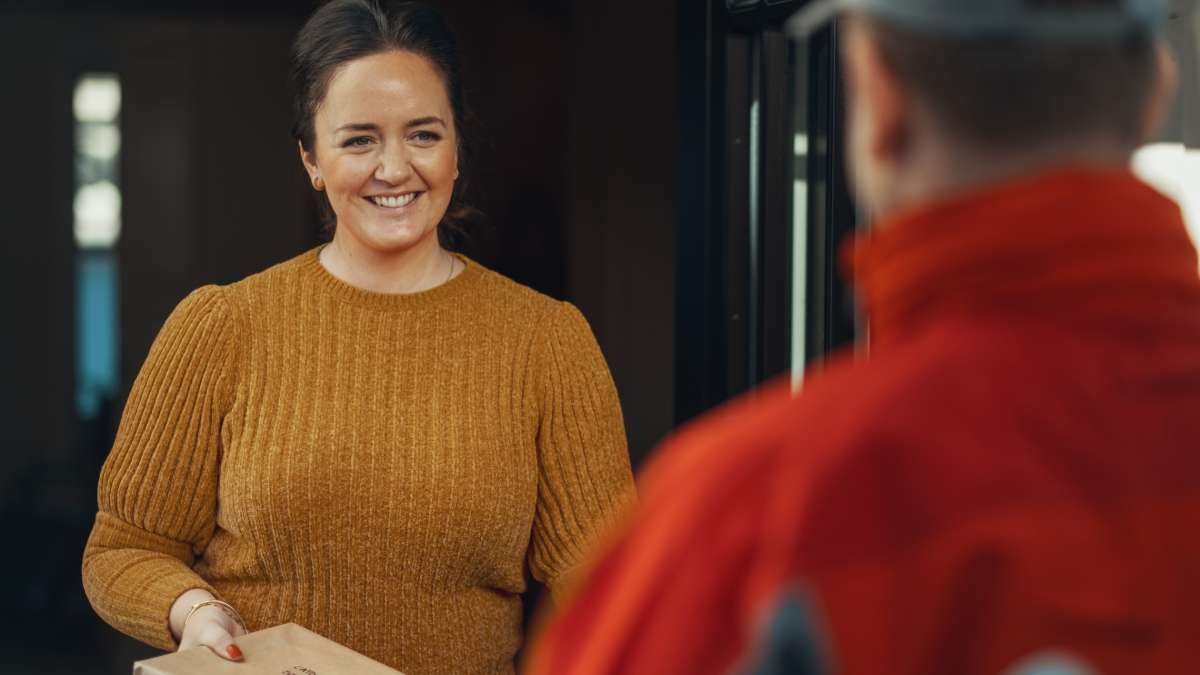 Smilende kvinne med pakke i hånden som hun rekker mot et postbud.