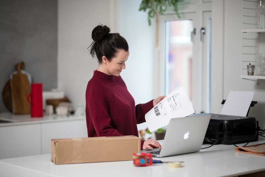 Dame med utskrift av adresselapp med tollinformasjon i hånda; og datamaskin og en printer på benken